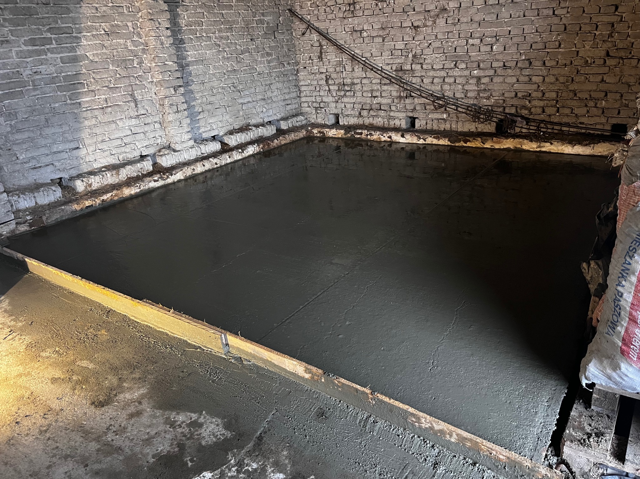 Świeżo wylana, mokra posadzka betonowa w pomieszczeniu z ceglanymi ścianami, widoczne drewniane szalunki i wystająca stal zbrojeniowa w górnej części ściany.