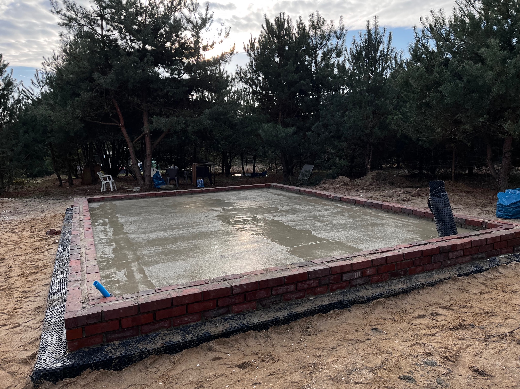 Świeżo wylana, mokra płyta betonowa otoczona czerwoną cegłą i czarną folią kubełkową na piaszczystym podłożu, otoczona sosnami.
