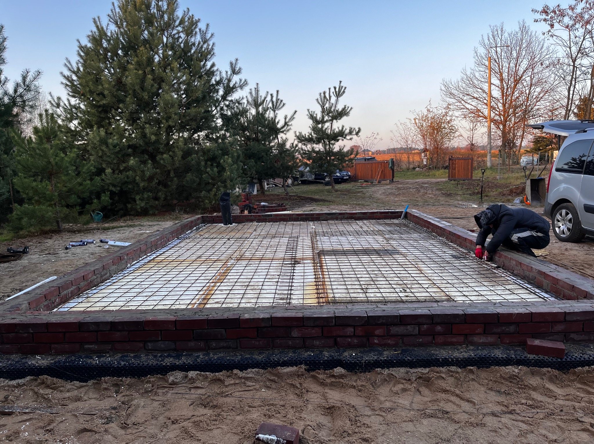 Przygotowanie zbrojenia na izolacji termicznej w fundamencie z cegły, widoczny pracownik w czerwonych rękawicach, otoczenie: piasek i drzewa.