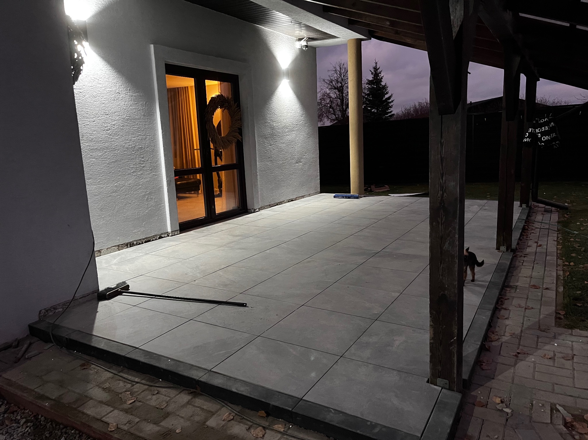 Wyłożony szarymi płytami taras przy domu z drewnianą konstrukcją zadaszenia, widoczne oświetlenie elewacyjne i wieniec na drzwiach wejściowych, mały pies w tle.