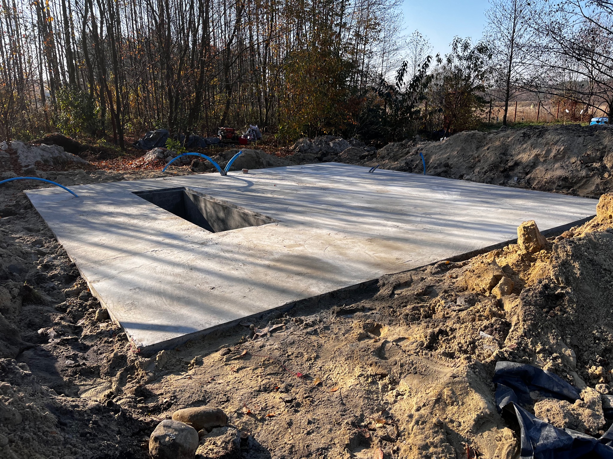 Świeżo wylana betonowa płyta fundamentowa z wycięciem na studzienkę, otoczona wykopaną ziemią i niebieskimi rurami, w tle drzewa bez liści w jesiennym słońcu.