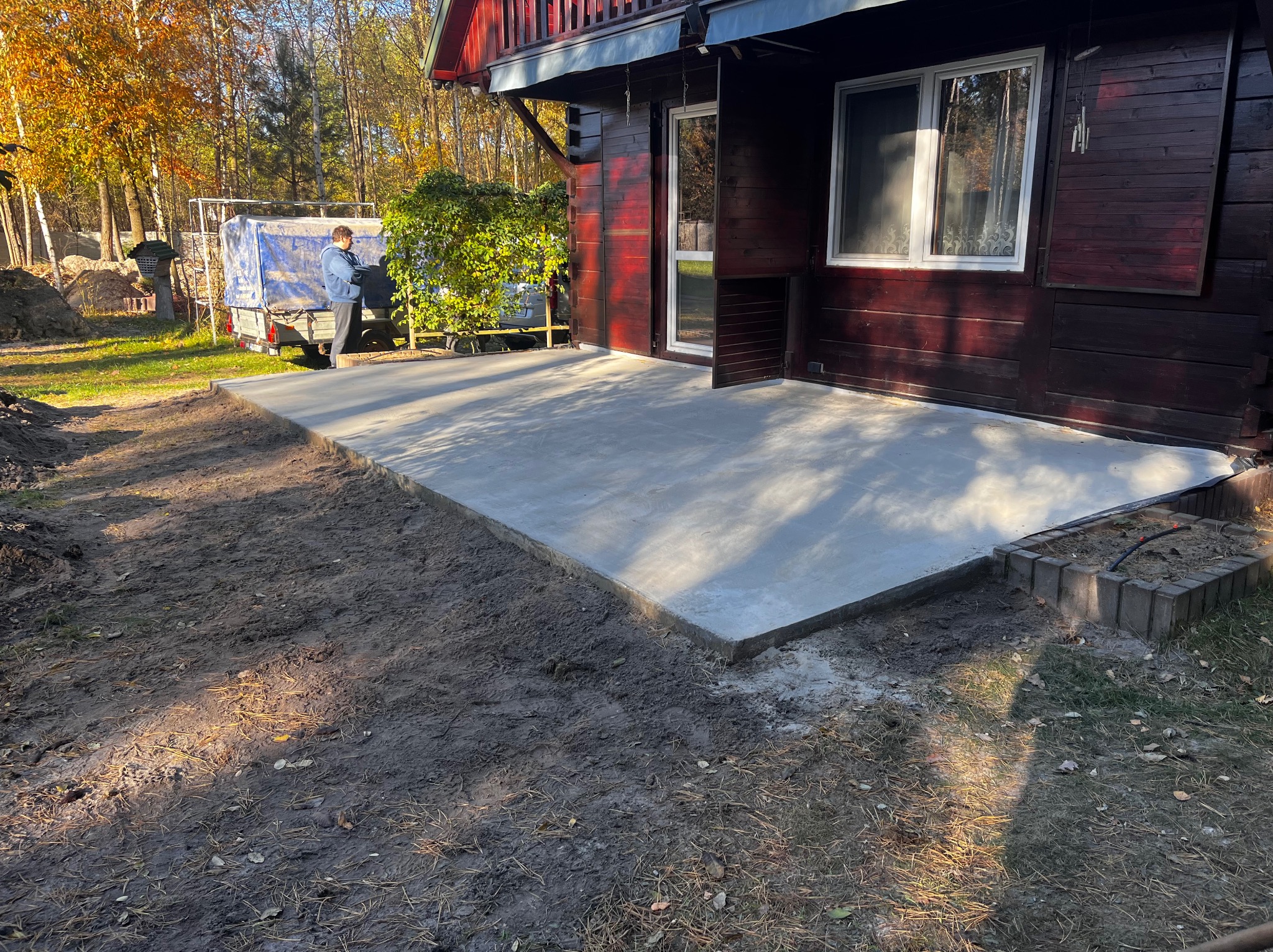 Świeżo wylany betonowy taras przylegający do drewnianego domu, widoczny fragment ogrodu i przyczepa z plandeką w tle.