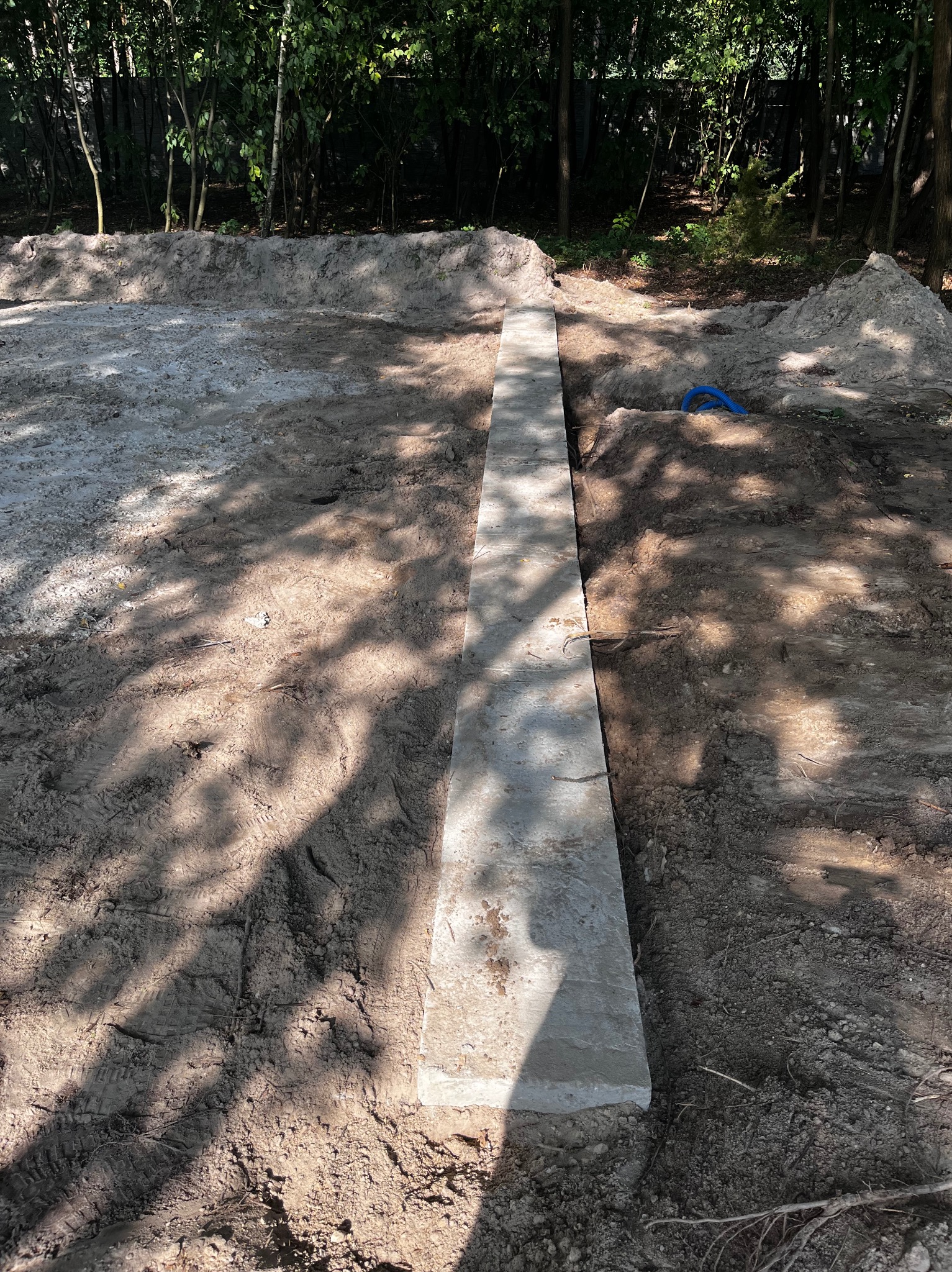 Przygotowany wykop pod fundament z ułożonym betonowym elementem szalunkowym i niebieską rurą w tle, widoczne ślady opon.