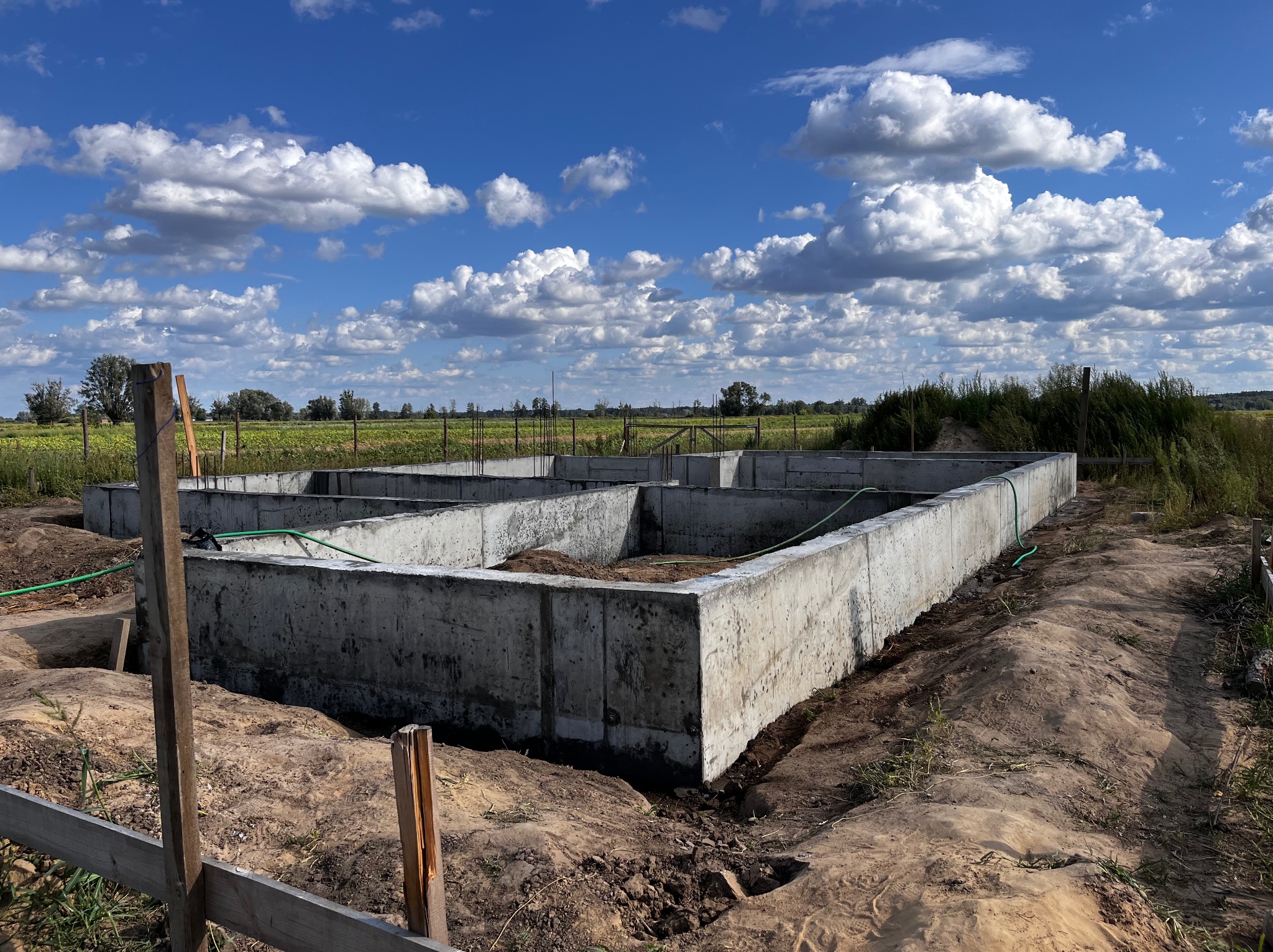 Betonowe ściany fundamentowe na etapie budowy, widoczne z zewnątrz, z zielonym wężem ogrodowym wewnątrz, na tle błękitnego nieba z białymi chmurami i zielonego pola.