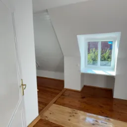 Pok&oacute;j na poddaszu w Ornecie po remoncie, z drewnianą podłogą częściowo odnowioną, białe ściany i sufit z oknem dachowym.