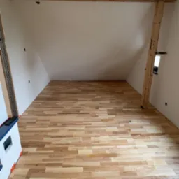 Jasna podłoga z paneli ułożona w skośnym pokoju na poddaszu. Widoczne drewniane belki stropowe i filar. Fragment grzejnika z boku kadru. Świeżo ułożone panele.