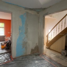 USŁUGI BUDOWLANE- OSKAR DULĘBA - Wnętrze w trakcie prac remontowych. Ściany przygotowane do szpachlowania, zabezpieczona podłoga folią. Widoczne schody w tle i narzędzia w sąsiednim pomieszczeniu.