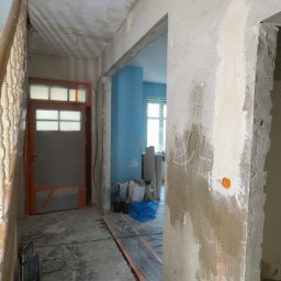 USŁUGI BUDOWLANE- OSKAR DULĘBA - Wnętrze w trakcie remontu: surowe ściany z widocznymi śladami szpachlowania, odkryta instalacja podłogowa, materiały budowlane w tle, przygotowanie do dalszych prac wykończeniowych.