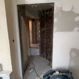 USŁUGI BUDOWLANE- OSKAR DULĘBA - Wnętrze w trakcie prac: ściany przygotowane pod gładzie, widoczne profile i izolacja. Na podłodze kabel i worek z odpadami budowlanymi.