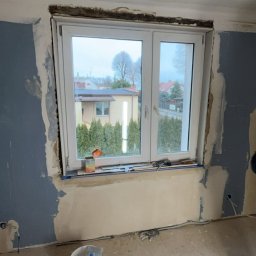 USŁUGI BUDOWLANE- OSKAR DULĘBA - Pokój w trakcie remontu: okno z widokiem na dom i tuje, ściany z częściowo nałożoną gładzią, wiadro z narzędziami na podłodze. Widoczne ślady po instalacji elektrycznej.