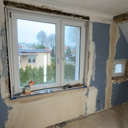USŁUGI BUDOWLANE- OSKAR DULĘBA - Wnętrze w trakcie remontu: okno z widokiem na zewnątrz, ściany częściowo pokryte gładzią, widoczne narzędzia i wiadro z zaprawą. Prace wykończeniowe w poddaszowym pokoju.