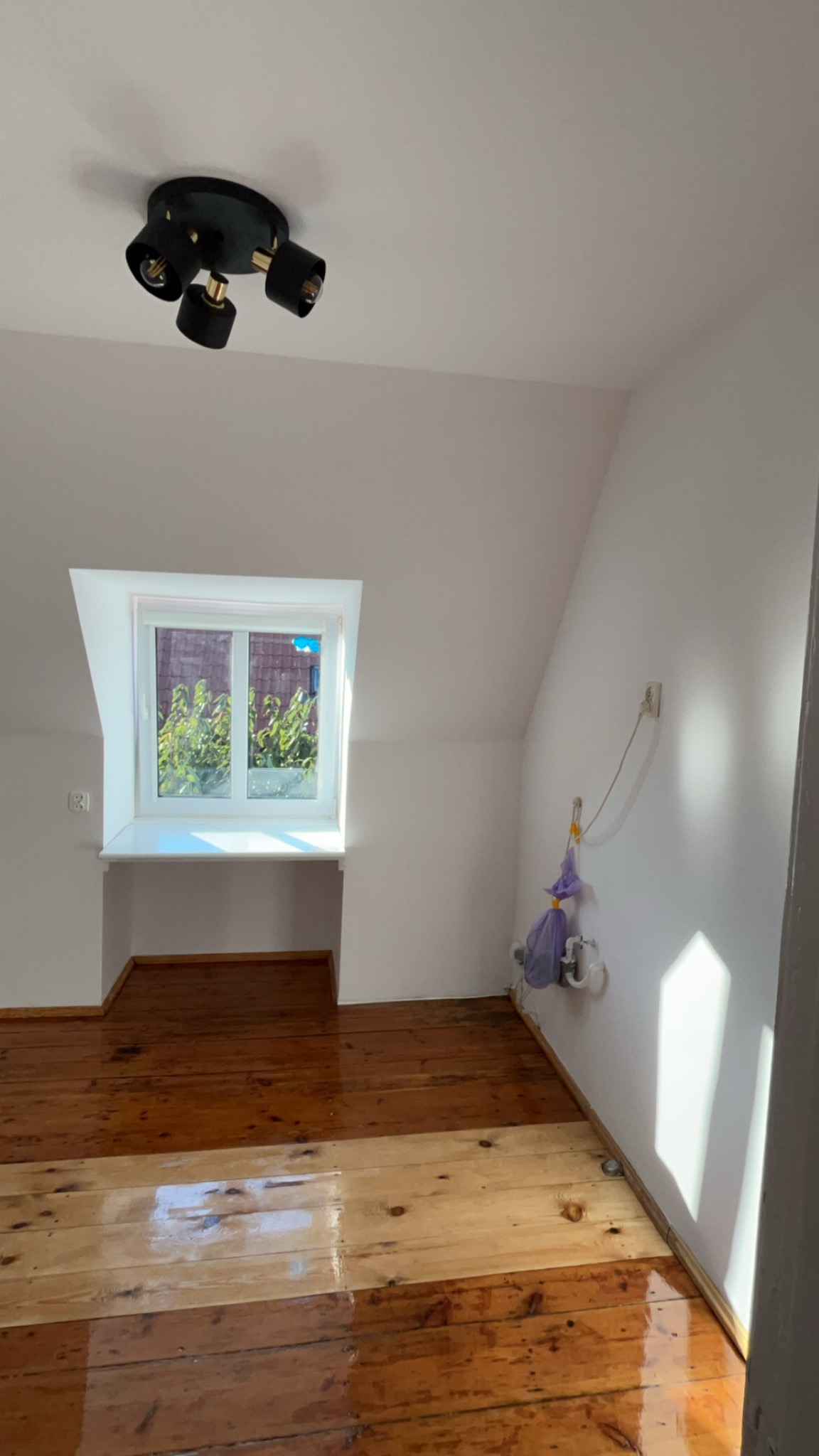 Pokój na poddaszu z drewnianą podłogą po cyklinowaniu, ściany pomalowane na biało, widoczna instalacja elektryczna przy ścianie i czarna lampa sufitowa.