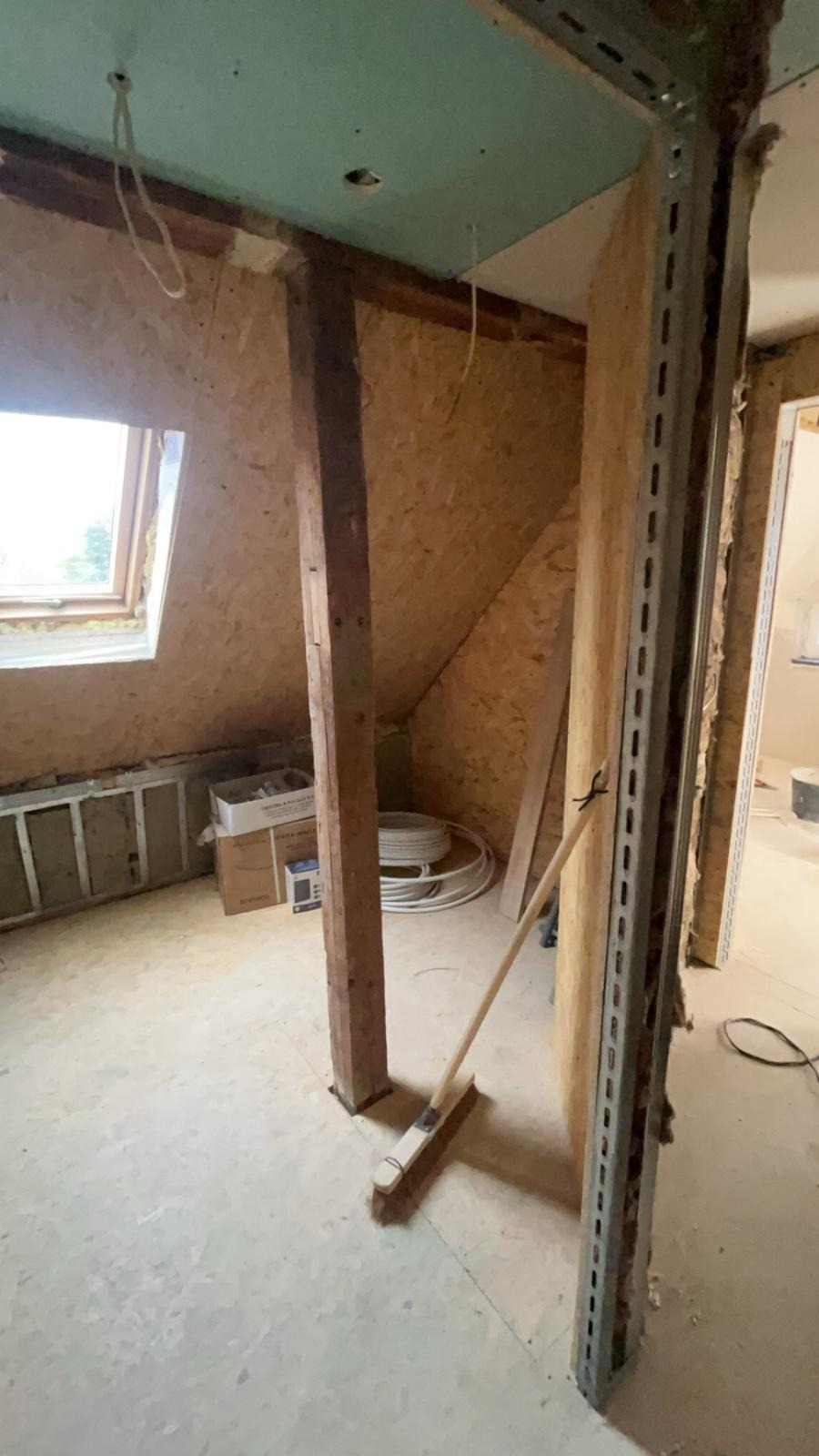 Poddasze w trakcie prac wykończeniowych: widoczne płyty OSB, stelaż metalowy, drewniany słup, okno dachowe i instalacja elektryczna. Przy ścianie oparta miotła.