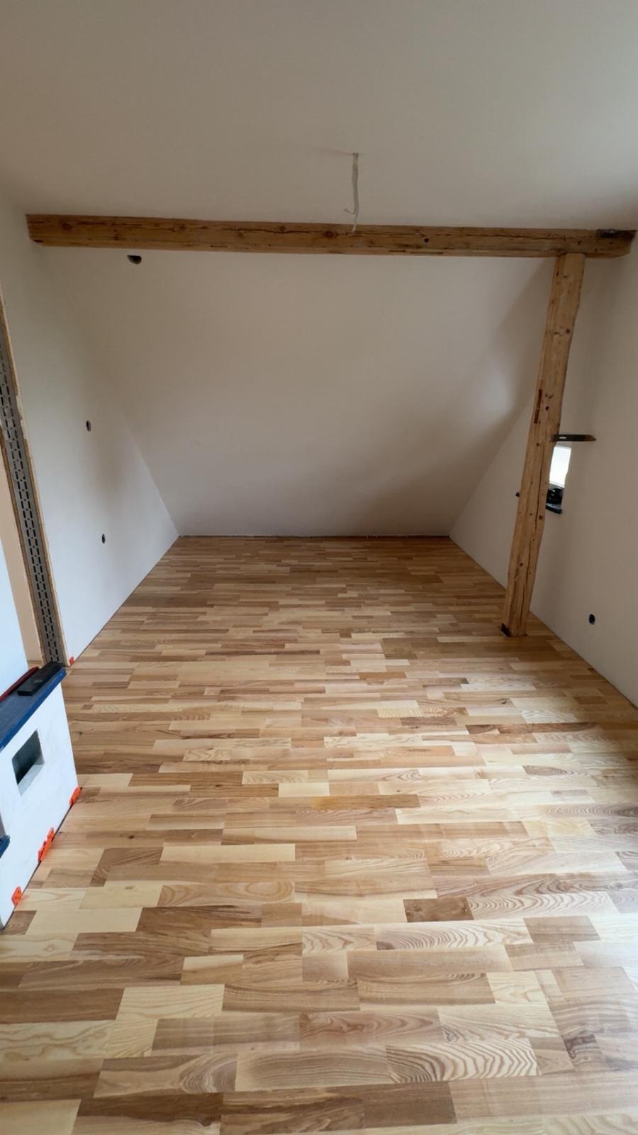 Jasna podłoga z paneli ułożona w skośnym pokoju na poddaszu. Widoczne drewniane belki stropowe i filar. Fragment grzejnika z boku kadru. Świeżo ułożone panele.