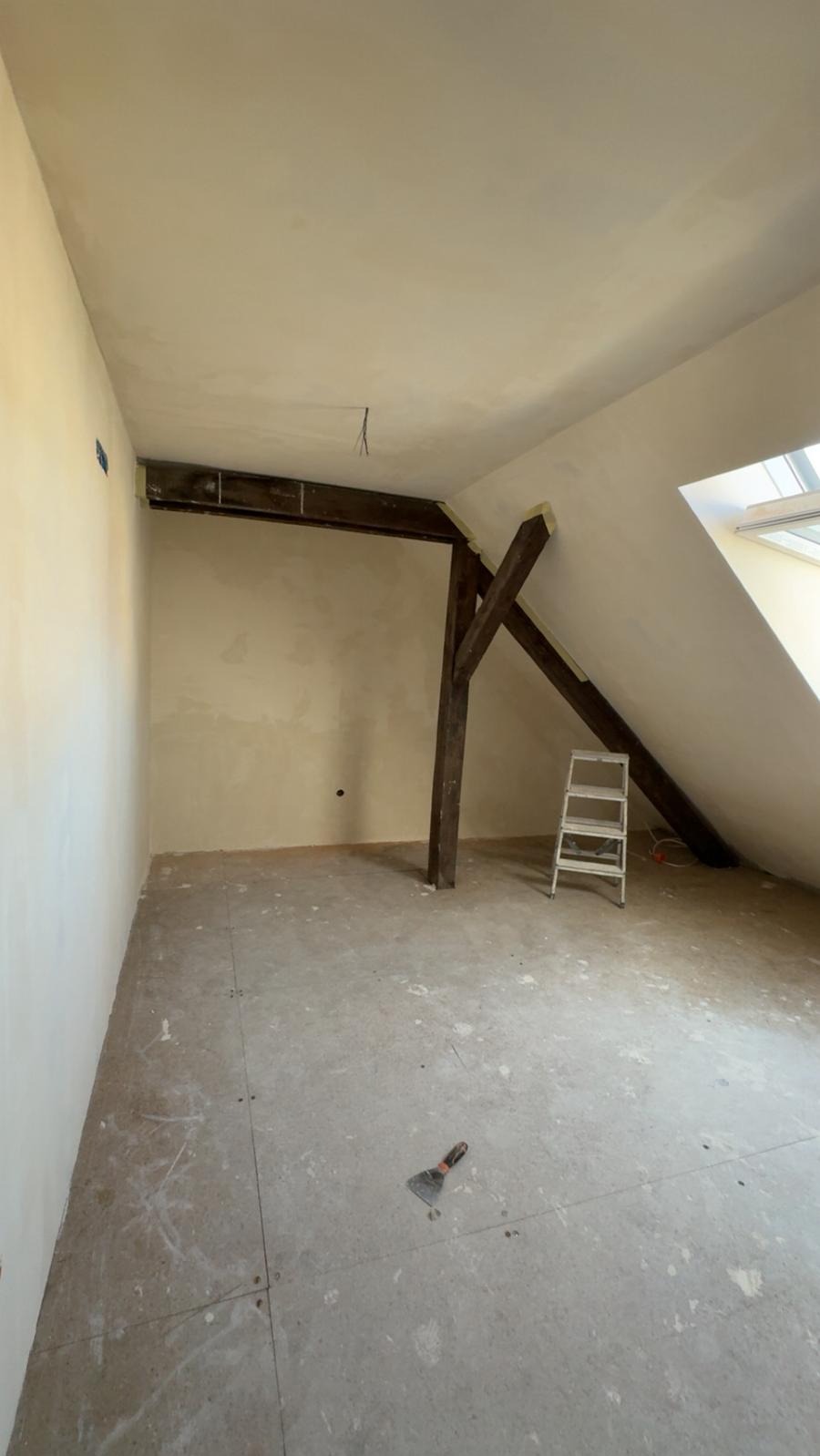 Poddasze w trakcie remontu: ściany po szpachlowaniu, widoczne drewniane elementy konstrukcyjne, drabina i szpachelka na podłodze. Przygotowanie do malowania.