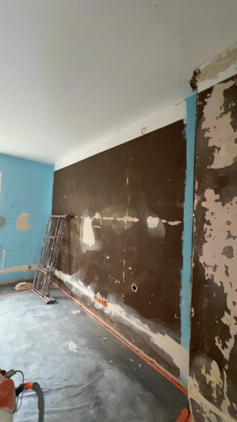Pokój w trakcie remontu: brązowa ściana ze śladami szpachli, drabina, podłoga zabezpieczona folią. Widoczne gniazdka elektryczne i fragment niebieskiej ściany.