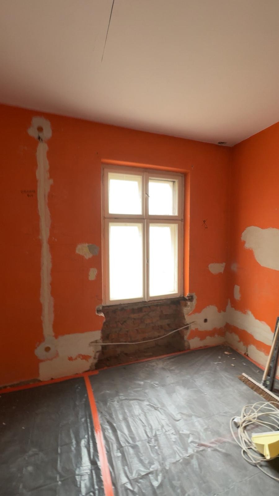 Pokój w remoncie: pomarańczowe ściany z widocznymi śladami szpachlowania, okno z odsłoniętą cegłą, podłoga zabezpieczona folią. Przygotowanie do dalszych prac.
