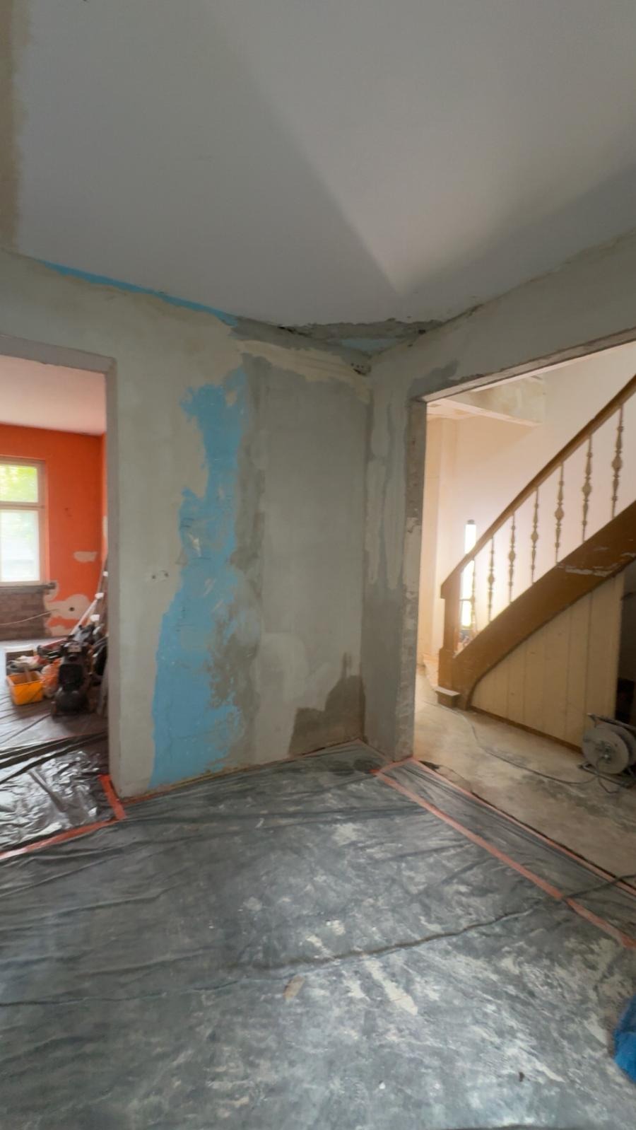 Wnętrze w trakcie prac remontowych. Ściany przygotowane do szpachlowania, zabezpieczona podłoga folią. Widoczne schody w tle i narzędzia w sąsiednim pomieszczeniu.