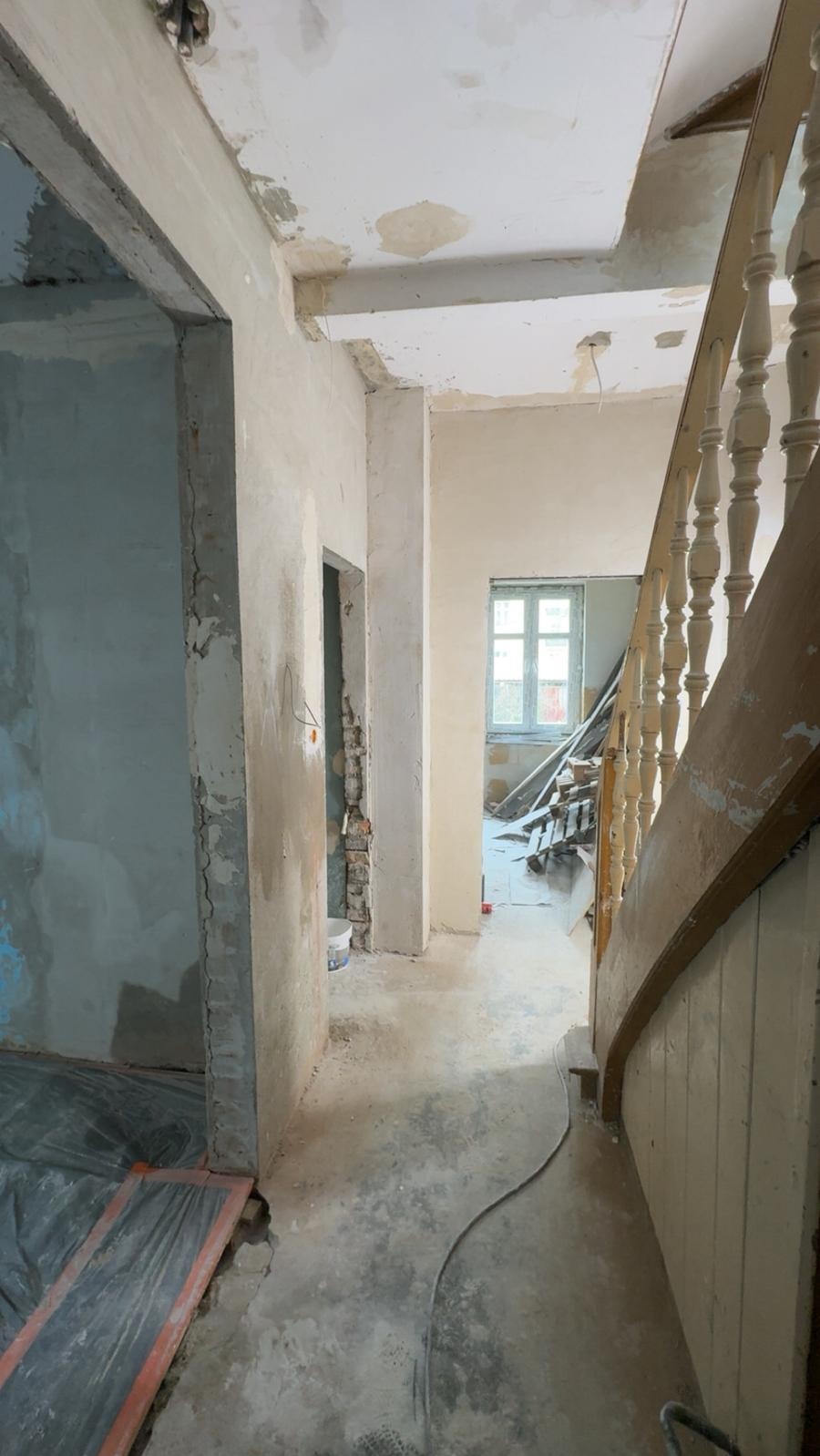 Wnętrze w trakcie remontu: przygotowane ściany do malowania, widoczne instalacje, podłoga zabezpieczona folią, schody z drewnianą balustradą. Prace wykończeniowe.