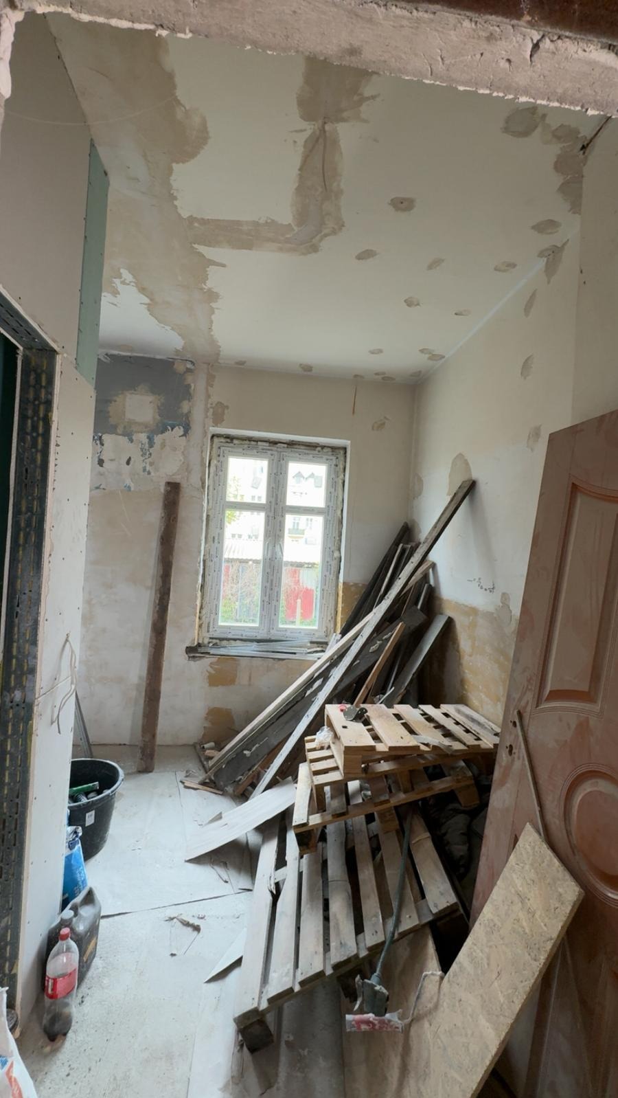 Wnętrze pokoju w trakcie remontu: ściany z ubytkami, częściowo zagruntowane, widoczne płyty karton-gips, okno, paleta i narzędzia malarskie. Przygotowanie do prac wykończeniowych.