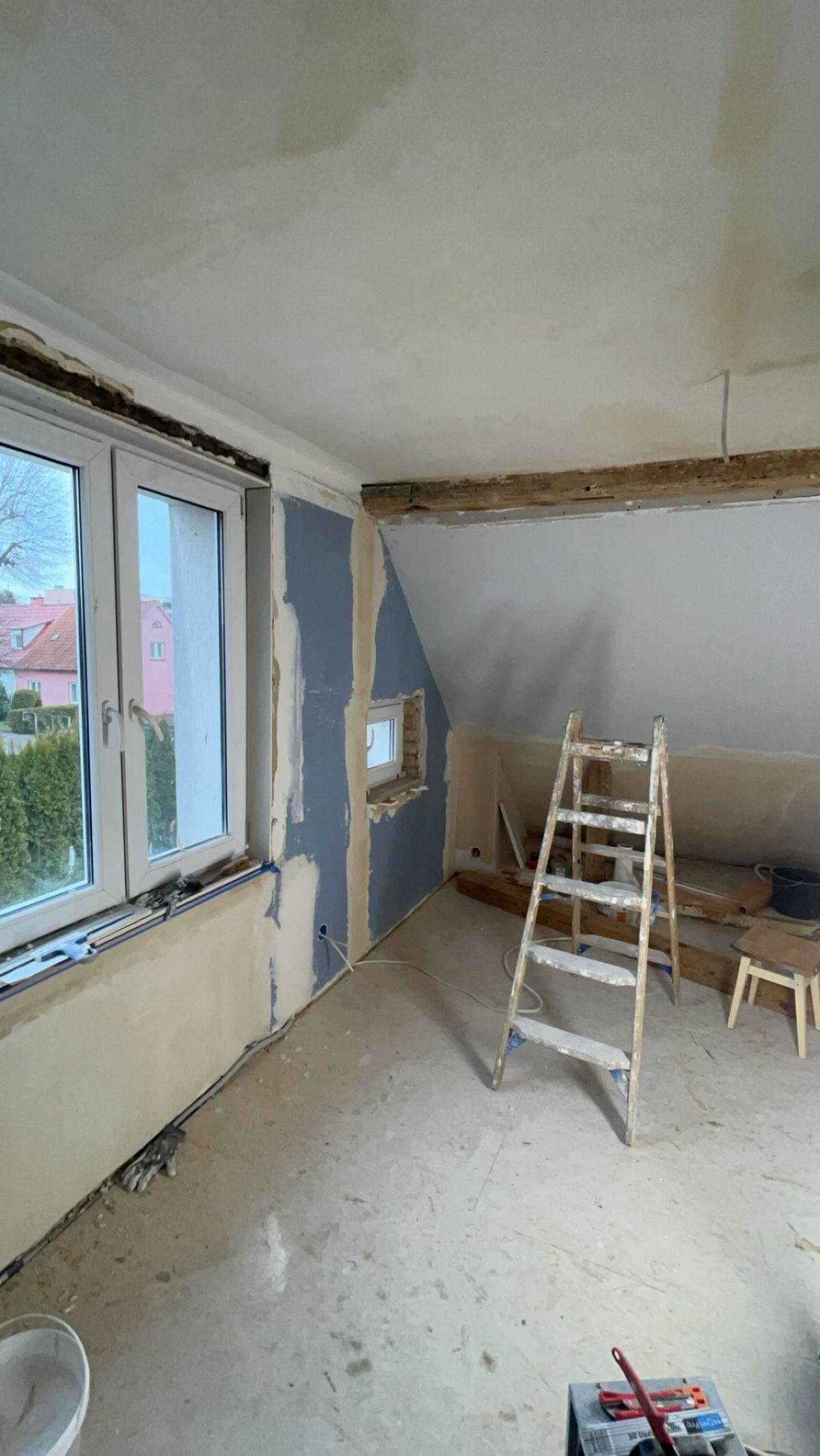 Poddasze w trakcie prac wykończeniowych: ściany częściowo wyszpachlowane, drabina malarska, okno dachowe, widoczne elementy drewniane konstrukcji.