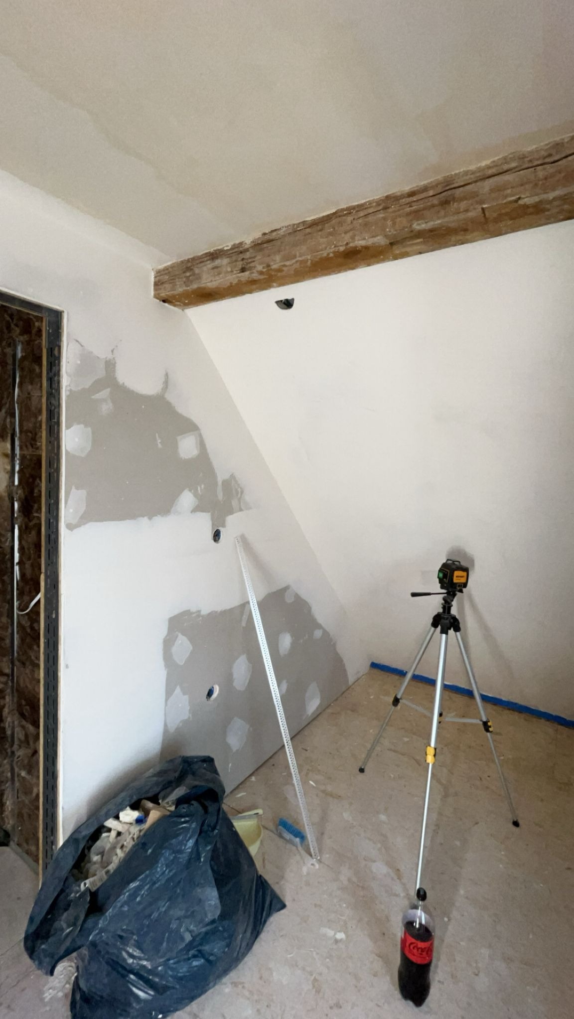 Poddasze w trakcie szpachlowania: widoczne fragmenty ścian z nałożoną gładzią, laser krzyżowy na statywie, worek z odpadami i drewniany element konstrukcyjny.