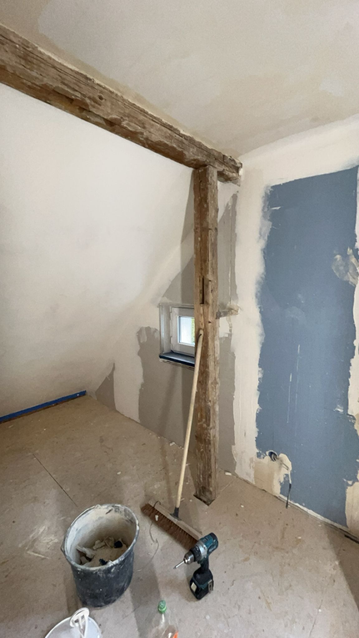 Poddasze w trakcie prac wykończeniowych: surowe drewniane belki, fragment ściany pomalowany na niebiesko, narzędzia i wiadro z zaprawą na podłodze.