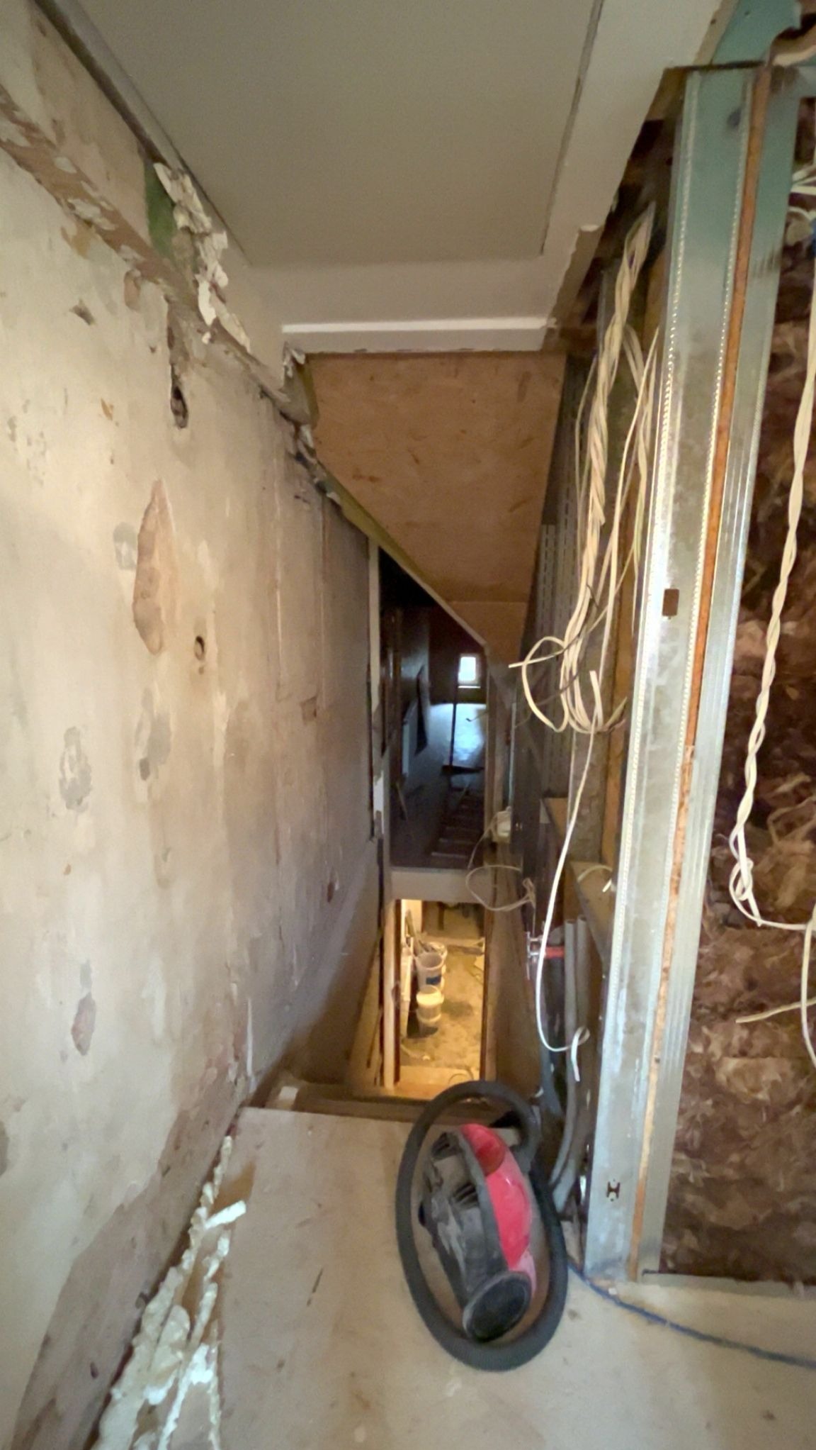 Klatka schodowa w trakcie remontu: odsłonięte ściany, metalowa konstrukcja, przewody elektryczne i odkurzacz na podłodze. Widoczne ślady po starych powłokach.