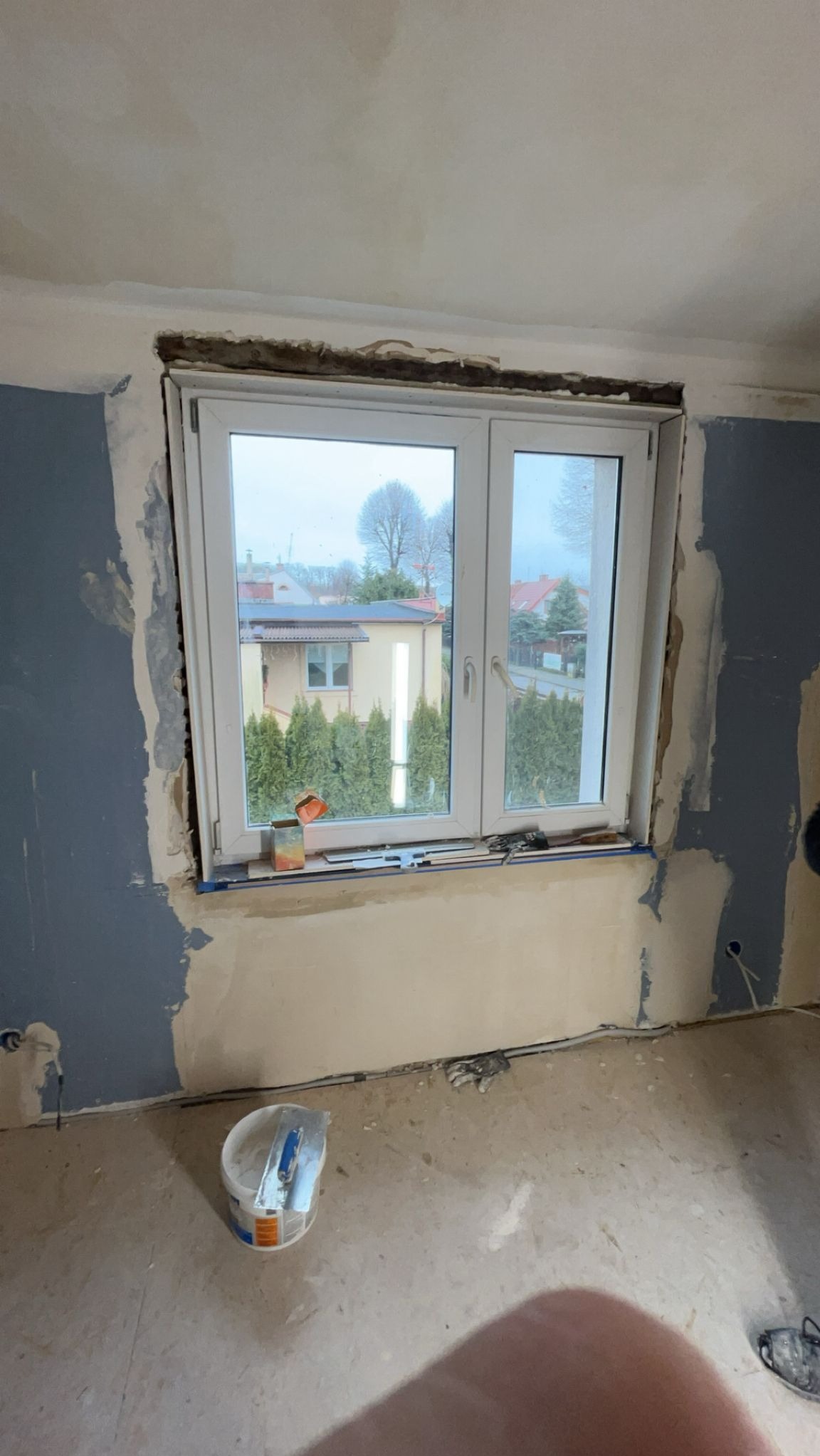 Pokój w trakcie remontu: okno z widokiem na dom i tuje, ściany z częściowo nałożoną gładzią, wiadro z narzędziami na podłodze. Widoczne ślady po instalacji elektrycznej.