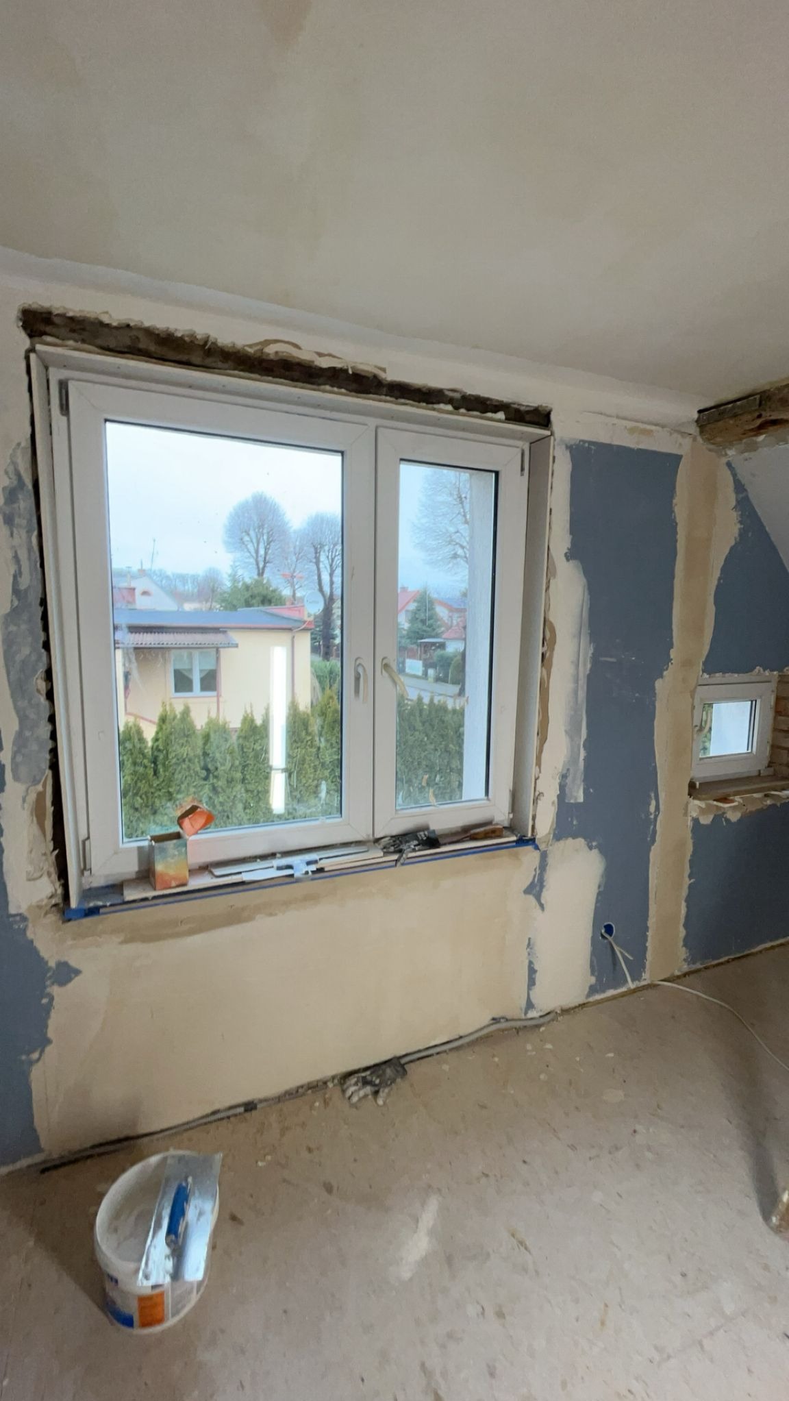 Wnętrze w trakcie remontu: okno z widokiem na zewnątrz, ściany częściowo pokryte gładzią, widoczne narzędzia i wiadro z zaprawą. Prace wykończeniowe w poddaszowym pokoju.
