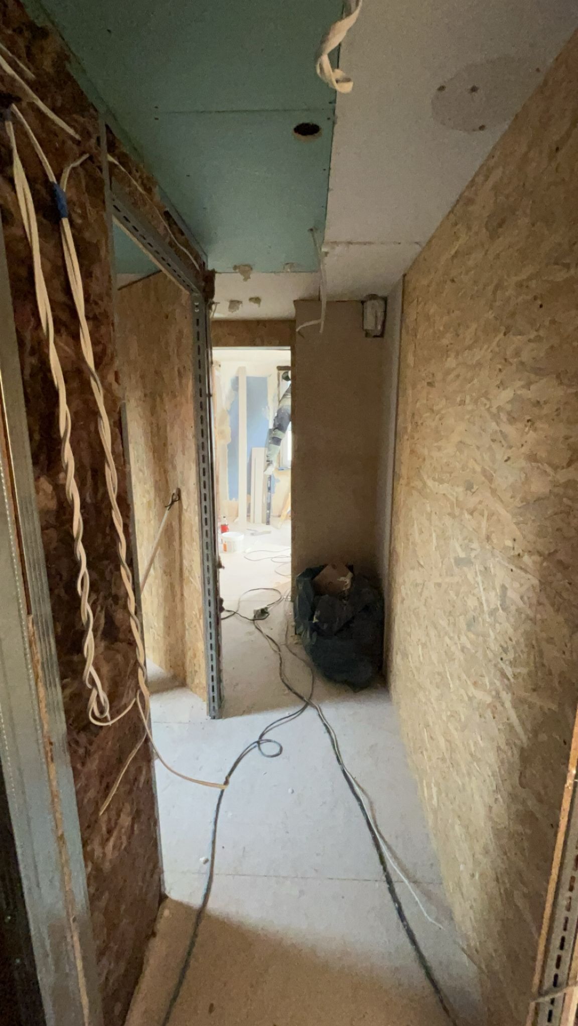 Korytarz w trakcie prac wykończeniowych: widoczne płyty karton-gips na suficie, przewody elektryczne i metalowe profile. W tle widok na kolejne pomieszczenie.