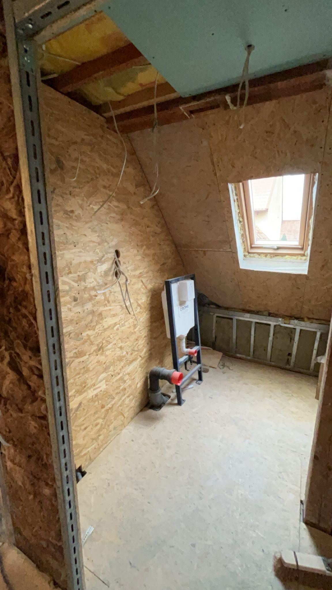 Adaptacja poddasza: montaż stelaża WC na tle ścian z płyty OSB, widoczne przewody instalacyjne i okno dachowe. Prace wykończeniowe w trakcie.