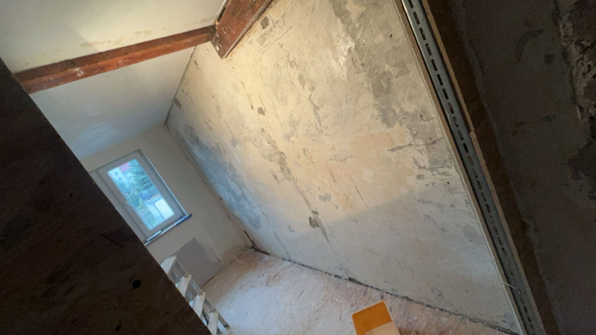 Wnętrze poddasza w trakcie remontu: surowe ściany z widocznymi śladami po wcześniejszych pracach, drewniane belki stropowe i fragment okna. Metalowa listwa na ścianie.