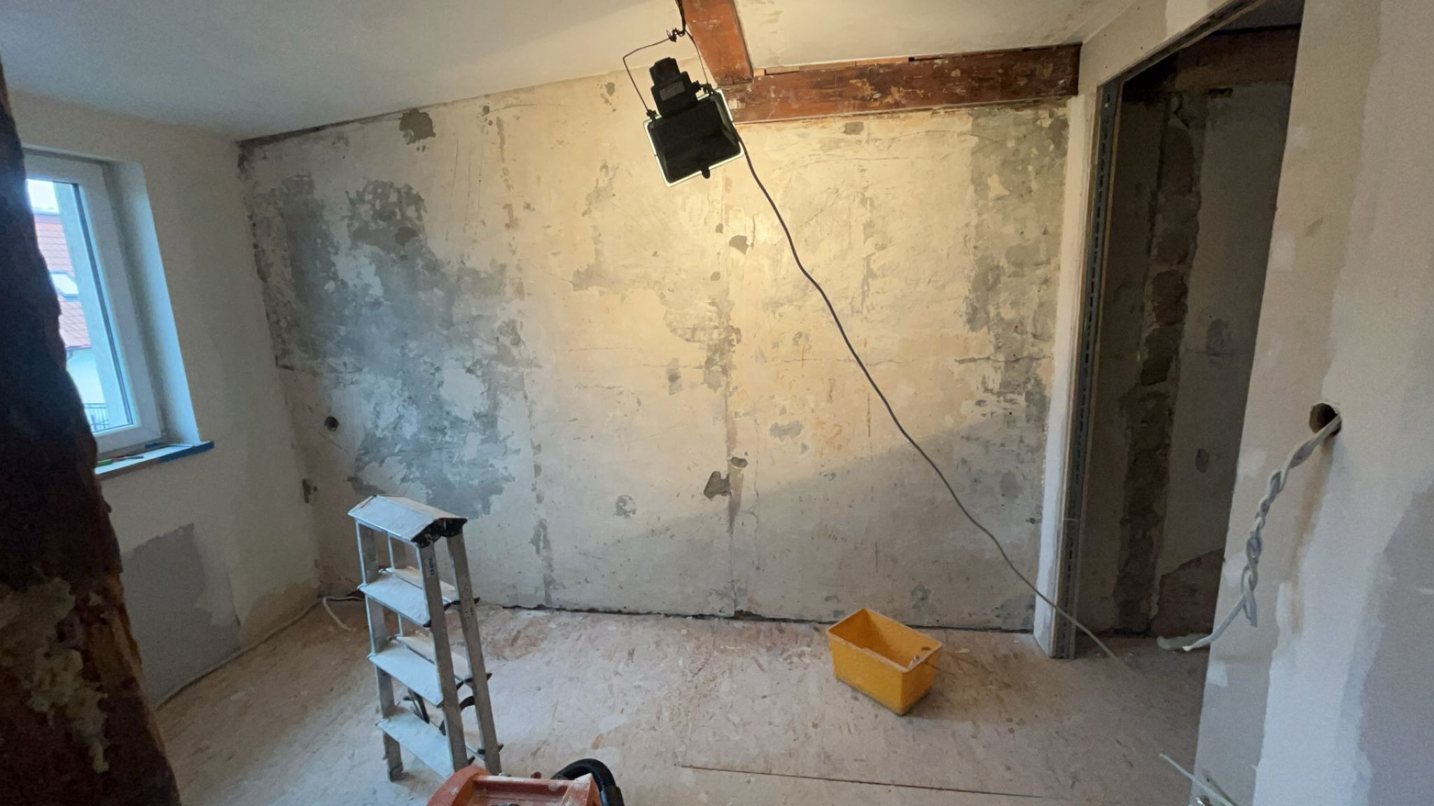 Pokój w trakcie remontu: ściany z widocznymi śladami po szpachlowaniu, drabina, wiadro i lampa halogenowa oświetlająca ścianę. Widoczny surowy stan wykończenia.
