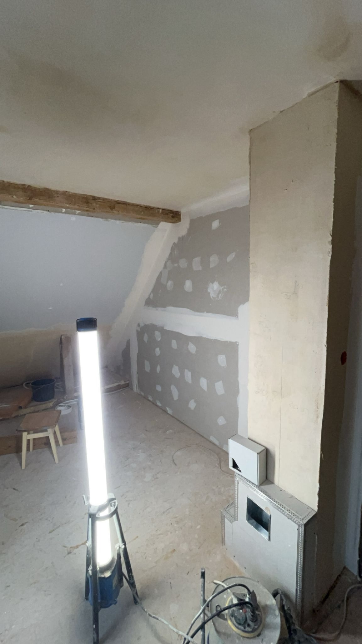 Pokój na poddaszu w trakcie prac wykończeniowych. Ściany z płyt gips-karton z widocznymi miejscami po szpachlowaniu, oświetlone lampą budowlaną, z elementami kominka.
