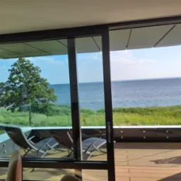 Przesuwne, szklane drzwi tarasowe w ciemnej ramie otwierają widok na drewniany taras z leżakami, za którym rozciąga się widok na morze i zieleń. Widoczny fragment wnętrza z krzesłem.