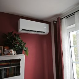 Klim-Tech Paweł Klimkiewicz - Biały klimatyzator ścienny z wyświetlaczem temperatury 21 stopni, zamontowany w pokoju z burgundową ścianą, obok okna z białymi zasłonami i półki z książkami i roślinami.