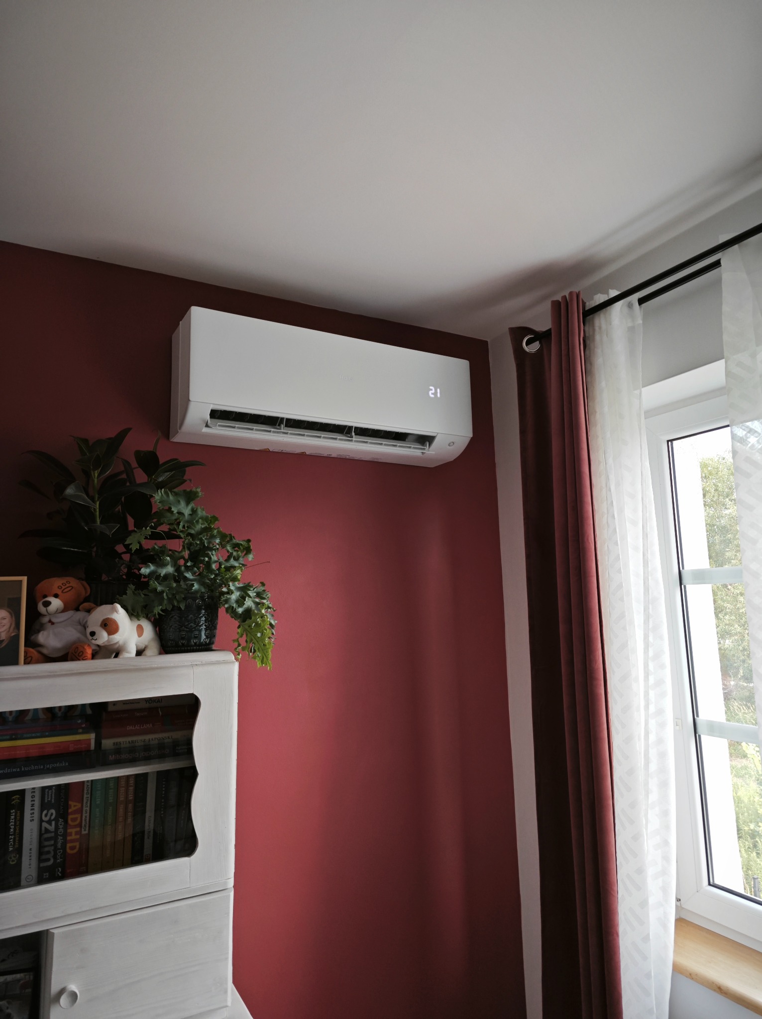 Biały klimatyzator ścienny z wyświetlaczem temperatury 21 stopni, zamontowany w pokoju z burgundową ścianą, obok okna z białymi zasłonami i półki z książkami i roślinami.