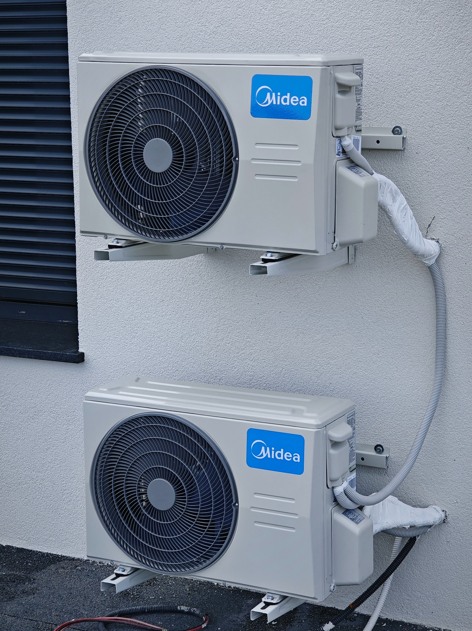 Dwa zewnętrzne moduły klimatyzacji Midea zamontowane na ścianie budynku z widoczną instalacją w białej osłonie. Dolny moduł umieszczony blisko gruntu.