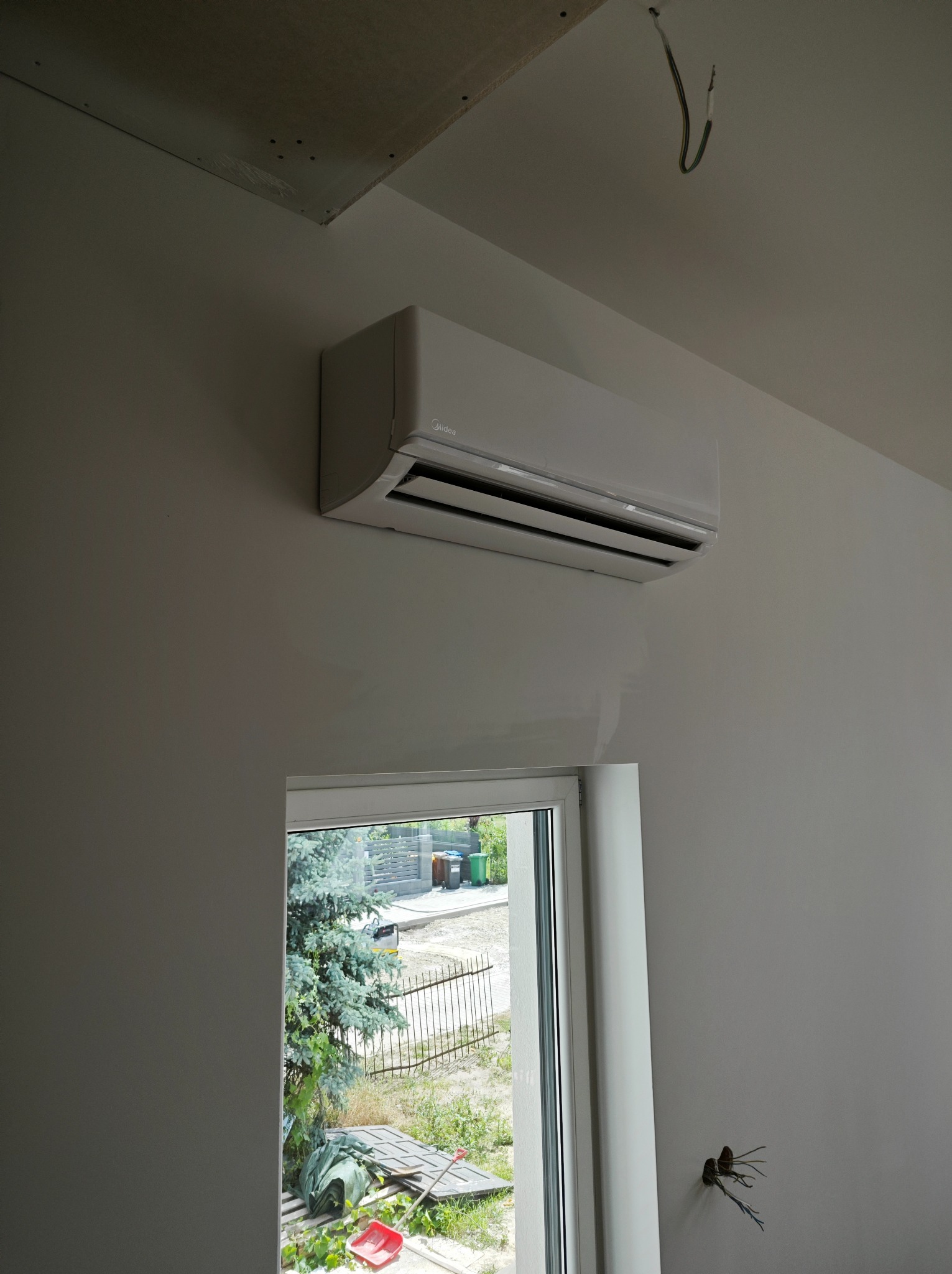 Biały klimatyzator Midea zamontowany na ścianie w pomieszczeniu w trakcie remontu, widoczne wystające kable i niedokończony sufit podwieszany.