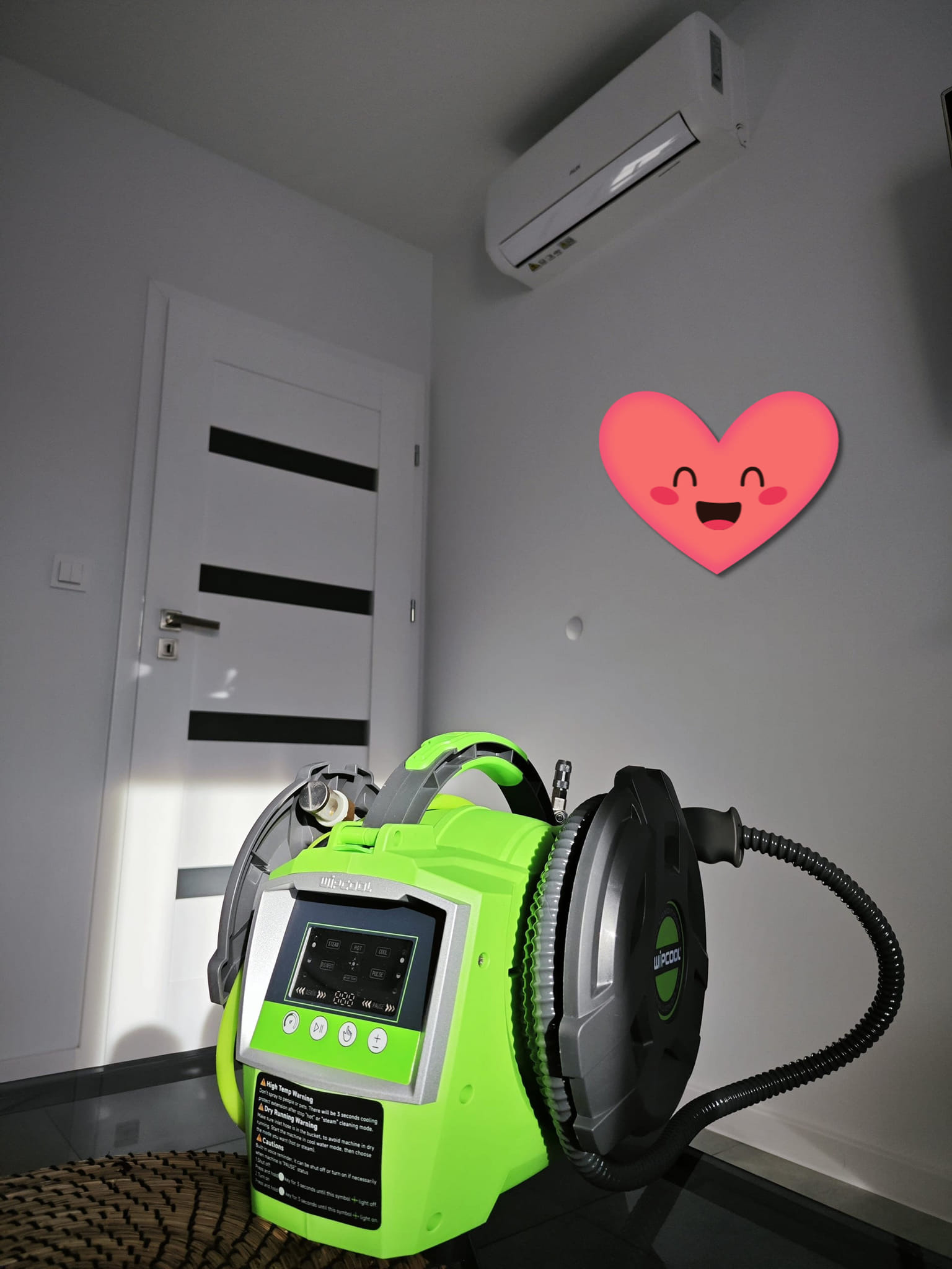 Myjka do klimatyzacji marki WIPCOOL z wyświetlaczem, w tle biały klimatyzator na ścianie i drzwi z czarnymi paskami, u góry naklejka w kształcie serca.