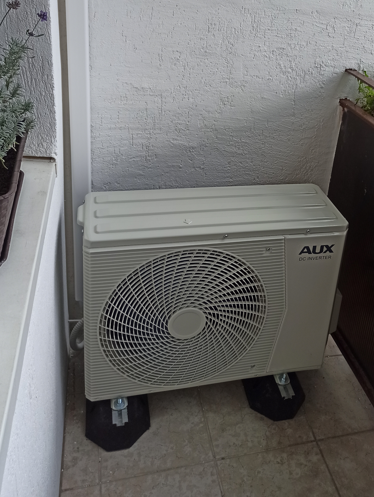 Zewnętrzna jednostka klimatyzacji AUX DC Inverter, zamontowana na balkonie na czarnych podkładkach antywibracyjnych, z widoczną teksturą ściany i roślinami w tle.