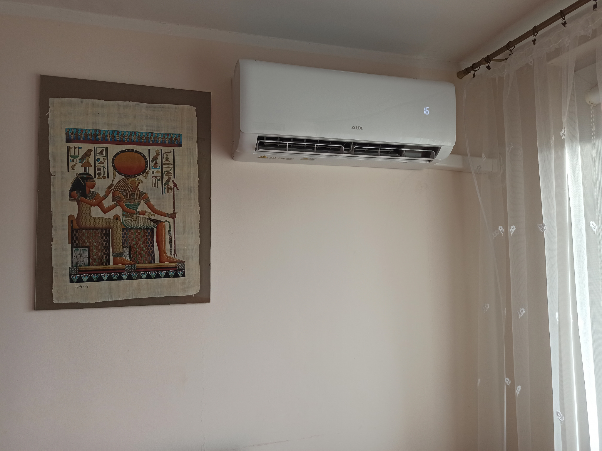 Biały klimatyzator AUX zamontowany na ścianie w pokoju, obok wisi obrazek z motywami egipskimi, a w oknie widać firankę.