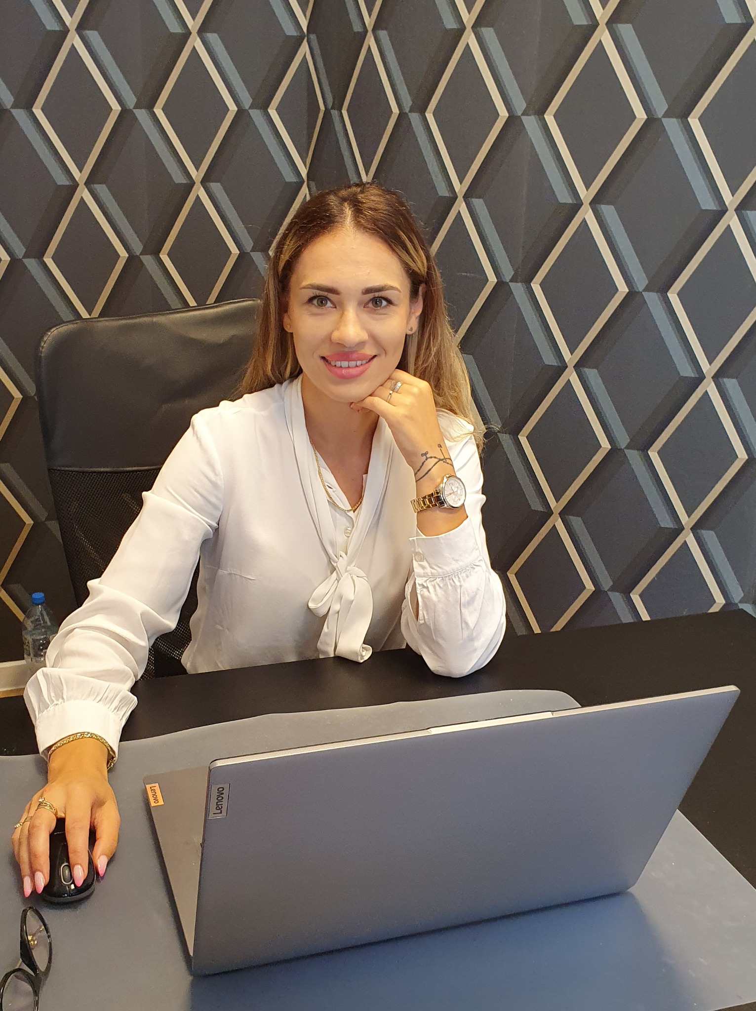Uśmiechnięta kobieta w białej bluzce siedzi za biurkiem z laptopem Lenovo, w tle geometryczna tapeta. Widoczny zegarek i tatuaż na nadgarstku.