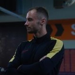 Mateusz Stańczyk TERNINGI PERSONALNE - Mężczyzna w czarnej bluzie sportowej z żółtymi wstawkami, stojący bokiem z założonymi rękami na tle siłowni z widocznym sprzętem i logo. Ostry profil twarzy, krótkie włosy zaczesane do tyłu.