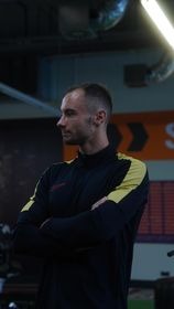 Mężczyzna w czarnej bluzie sportowej z żółtymi wstawkami, stojący bokiem z założonymi rękami na tle siłowni z widocznym sprzętem i logo. Ostry profil twarzy, krótkie włosy zaczesane do tyłu.