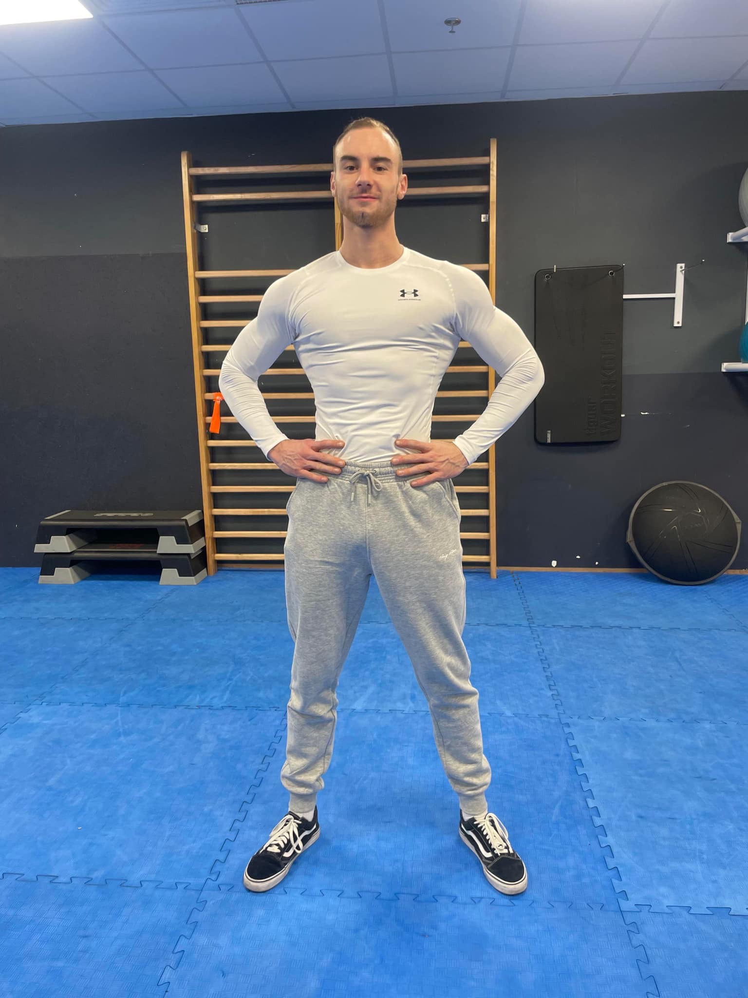 Mężczyzna w sportowym stroju, biała koszulka z długim rękawem i szare dresowe spodnie, stoi w siłowni na niebieskiej macie, ręce oparte na biodrach, w tle drabinki gimnastyczne i sprzęt do ćwiczeń.