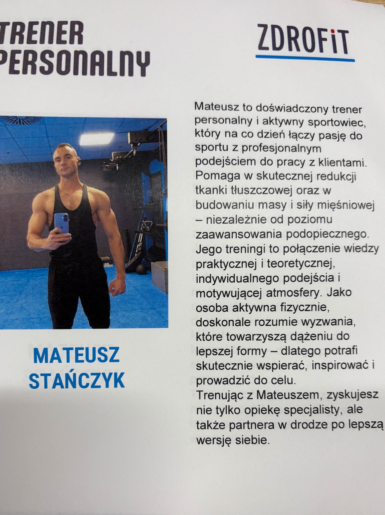 Przystojny trener personalny z Pruszkowa prezentuje sylwetkę na tle siłowni, trzymając telefon. Widoczny napis 'Trener Personalny' i logo 'Zdrofit'.