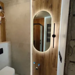 Nowoczesna łazienka z podświetlanym lustrem owalnym, drewnopodobne płytki na ścianie, wisząca toaleta i widoczny fragment kabiny prysznicowej.