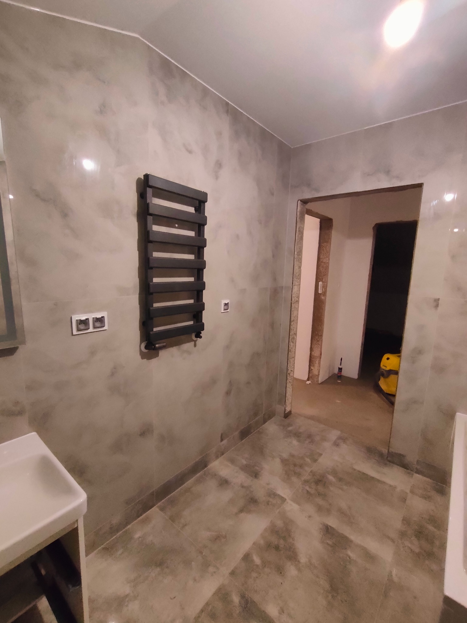 Wykończona łazienka z szarymi płytkami imitującymi kamień, grzejnikiem drabinkowym na ścianie, widoczną umywalką i otwartym przejściem do innego pomieszczenia w trakcie prac.